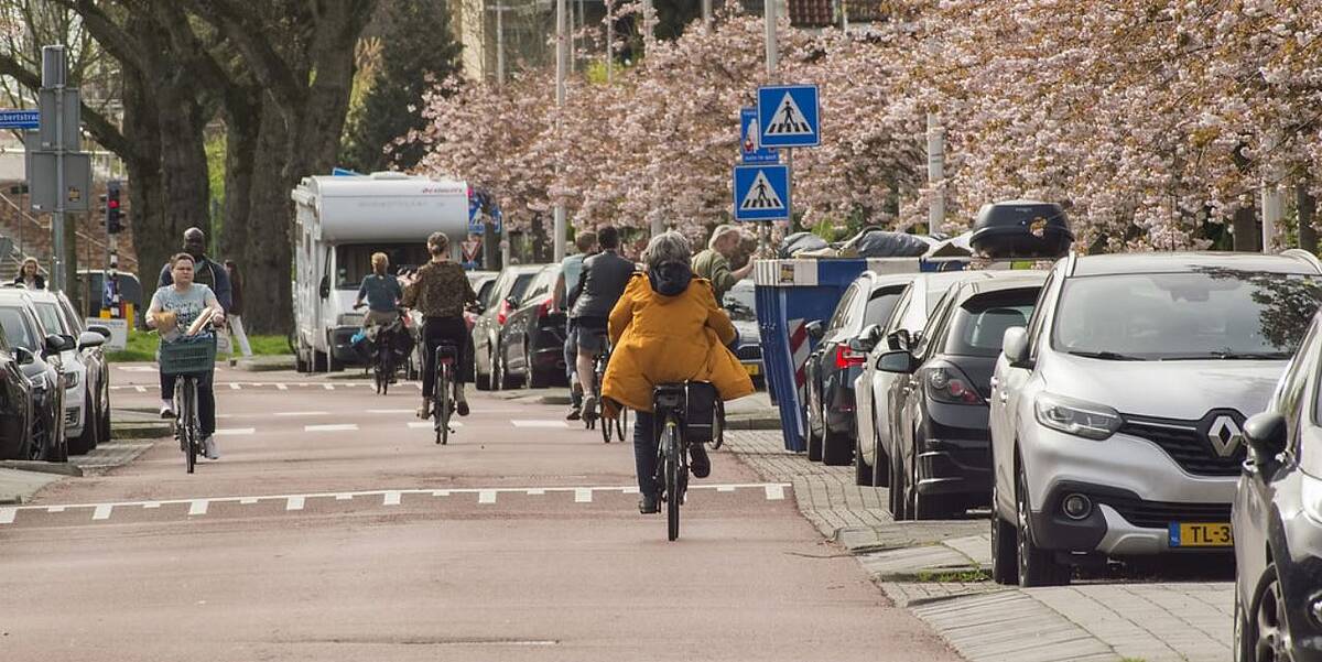 Link naar foto in Flickr: fietsers in de straat. Aan biede kanten geparkeerde auto's en bloesem.