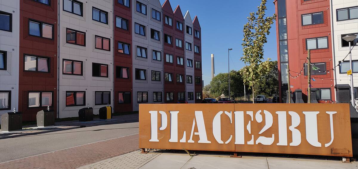 klik en vergroot Place2BU bord met op de achtergrond woningen