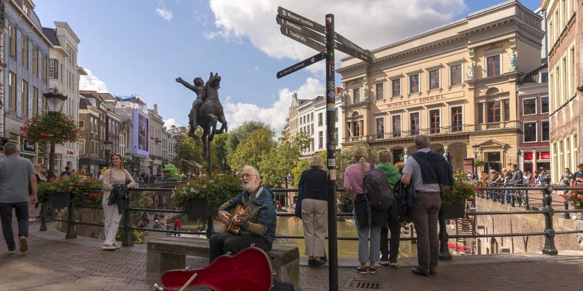Link naar foto in Flickr: mensen staan langs de railing bij de gracht op de Stadhuisbrug. Op de voorgrond een gitaarspeler op een bankje.