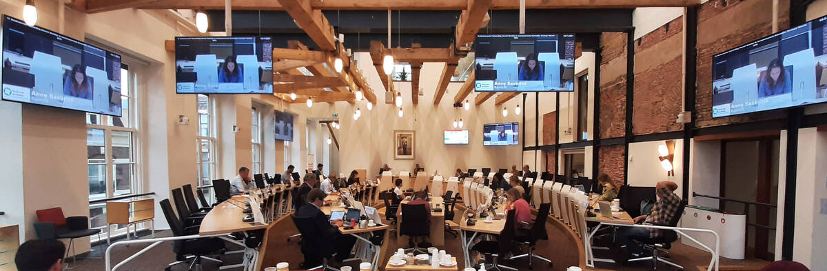 Foto van de Utrechtse raadszaal, waar de raad aan het vergaderen is. Er zijn schermen te zien waarop de livestream van de vergadering zichtbaar is.