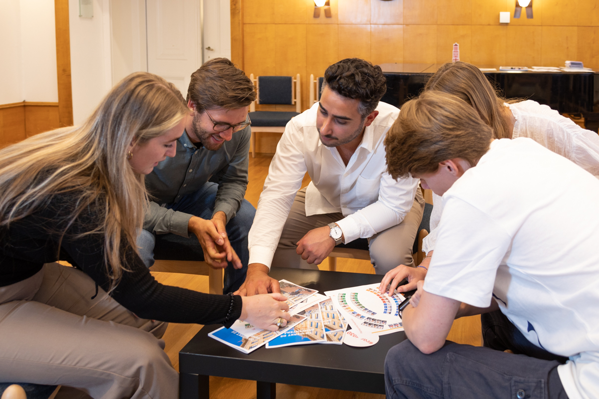 Foto van een groep mensen die samen tijdens een cursus naar materiaal over de gemeenteraad kijkt. De groep zit op stoelen in een kring om een lage tafel heen.