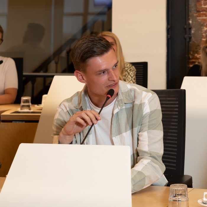 Jongere in de raadszaal van het stadhuis, die door de microfoon spreekt en op de stoel van een raadslid zit.